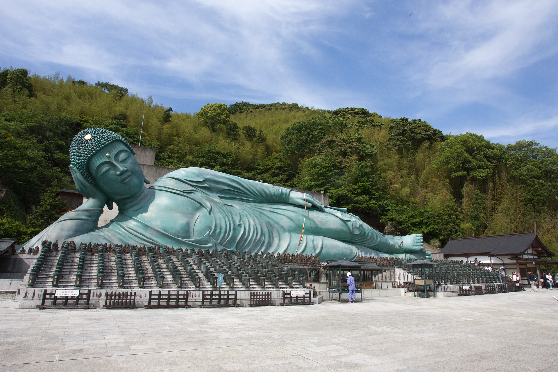 Wide view of the Nanzoin Reclining Buddha with hundreds of small standing Buddha statues lined up in front and the wooded hill behind