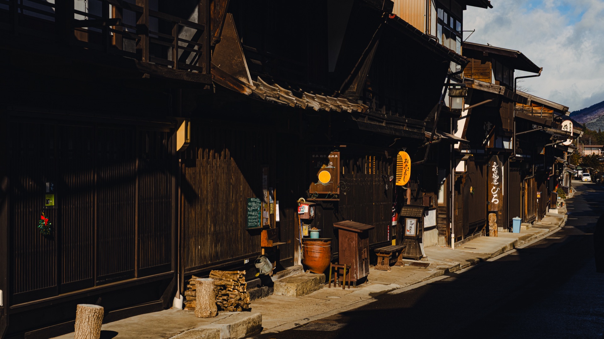 A traditional Naraijuku storefront in afternoon shadow with a yellow lantern noren curtain and stacked firewood