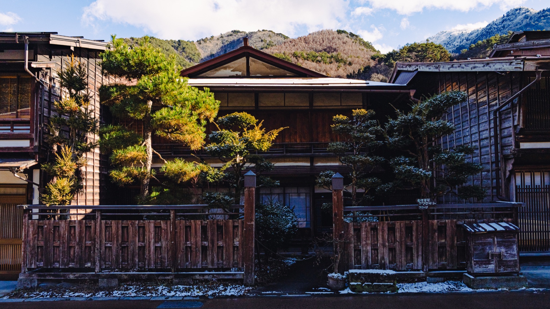 A traditional ryokan facade with bonsai pines and the Kiso mountains showing fresh snow behind