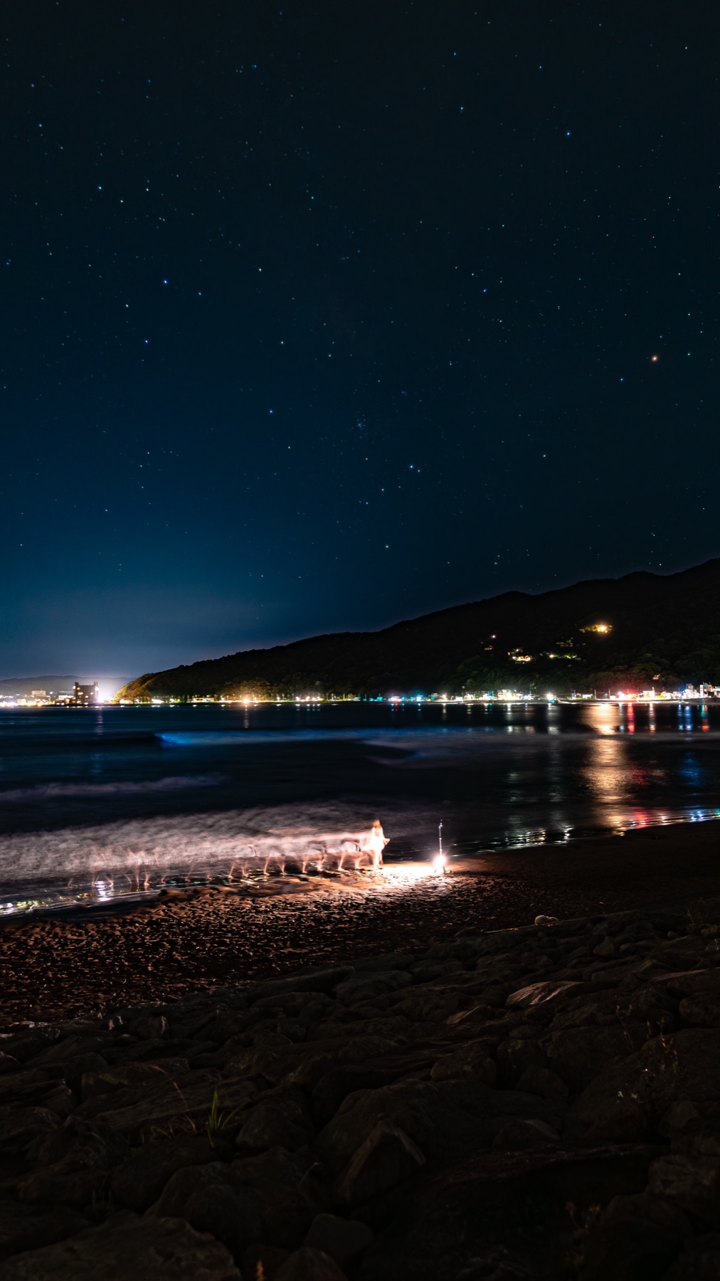Vertical night photo of a dark rocky beach with a warm light trail in the foreground and a faint blue line of bioluminescent waves, under a sky thick with stars.