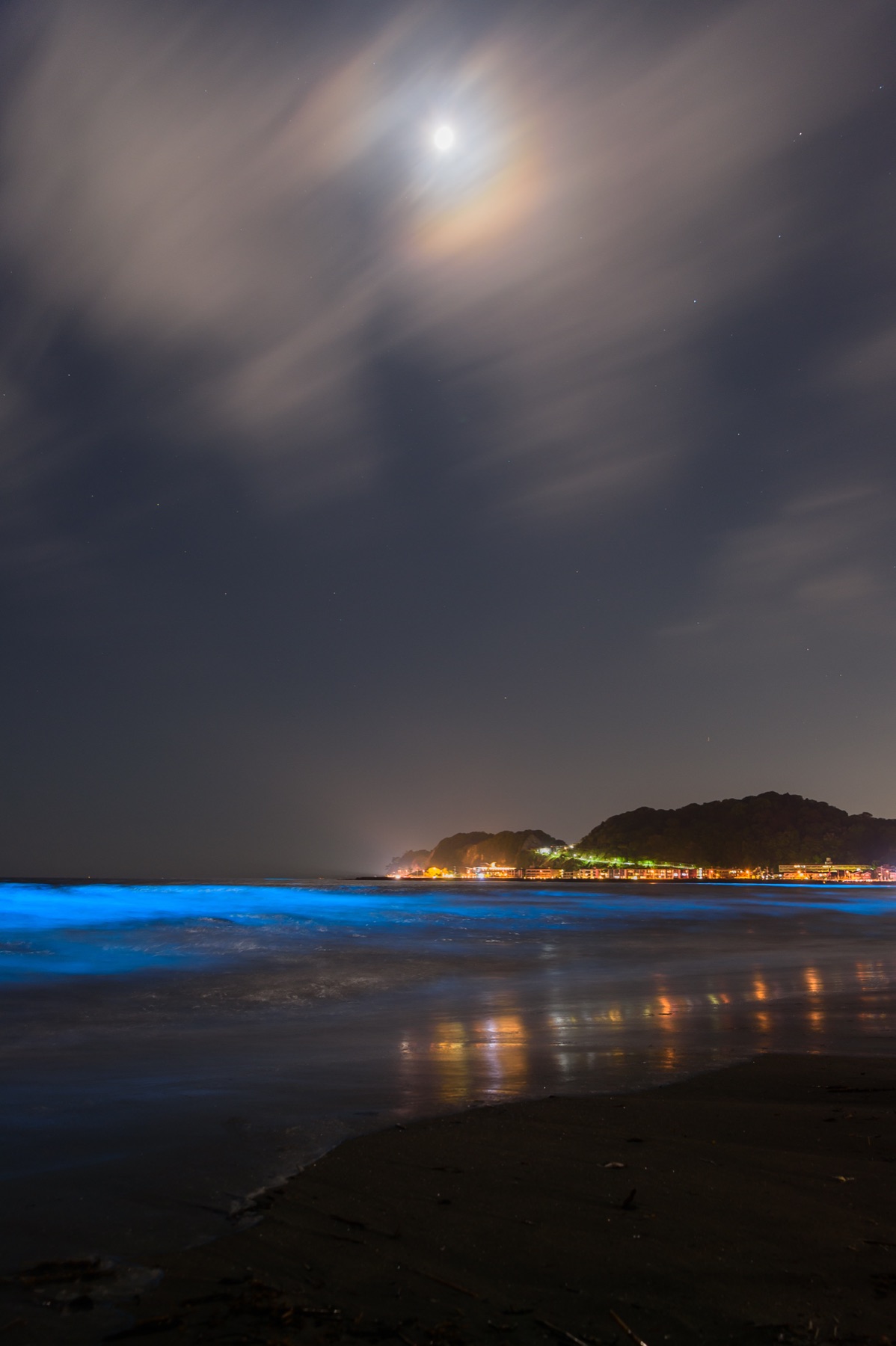 Vertical long-exposure photo of bioluminescent blue sea-sparkle waves washing onto a dark beach at night, with a bright moon breaking through clouds and a lit headland in the distance.