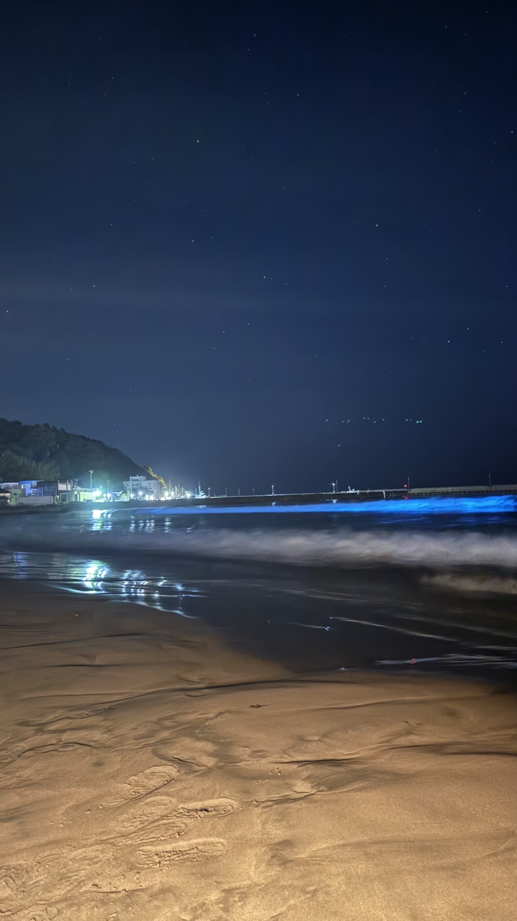 Smartphone night-mode photo of a beach where the breaking wave line glows vivid electric blue, with footprints in the wet sand foreground and town lights in the distance.