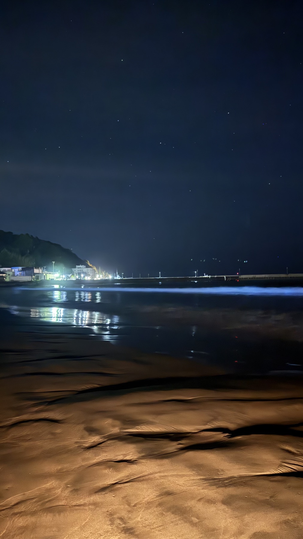 Smartphone photo of the same beach on a weaker night, where the bioluminescent wave line glows a softer fainter blue, with reflections of town lights on the wet sand.
