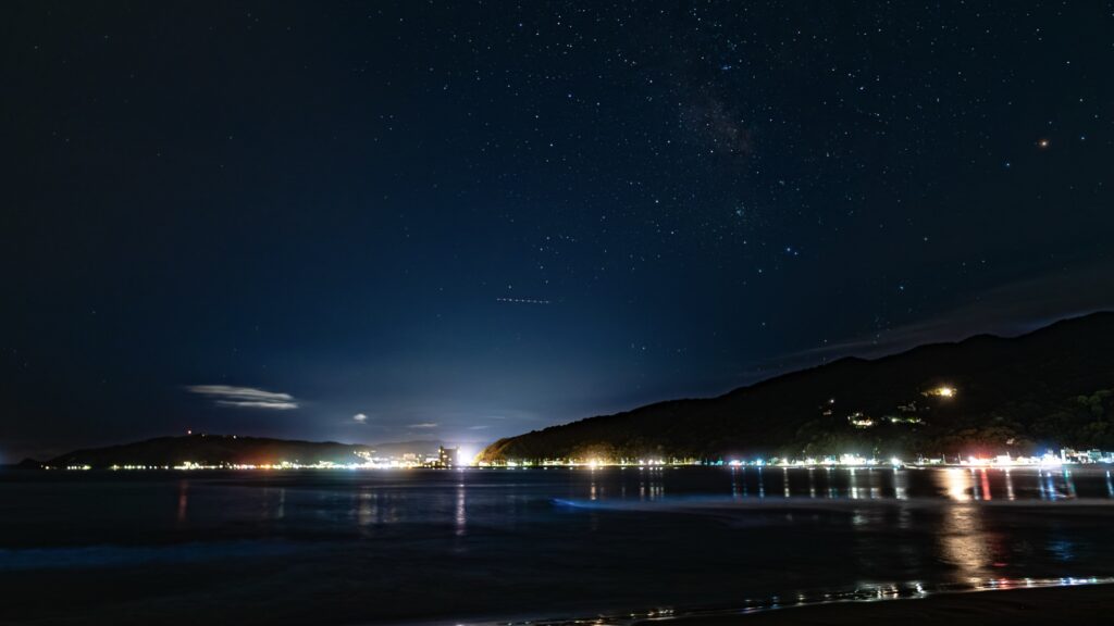 Wide long-exposure night photograph of a Japanese bay under a star-filled sky, with town lights along the far shore and a faint blue band of bioluminescent sea sparkle glowing at the wave line.