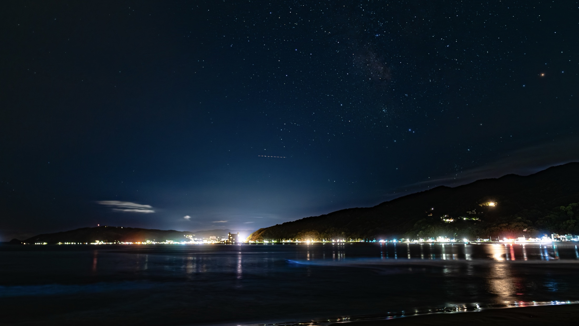 Wide long-exposure night photograph of a Japanese bay under a star-filled sky, with town lights along the far shore and a faint blue band of bioluminescent sea sparkle glowing at the wave line.
