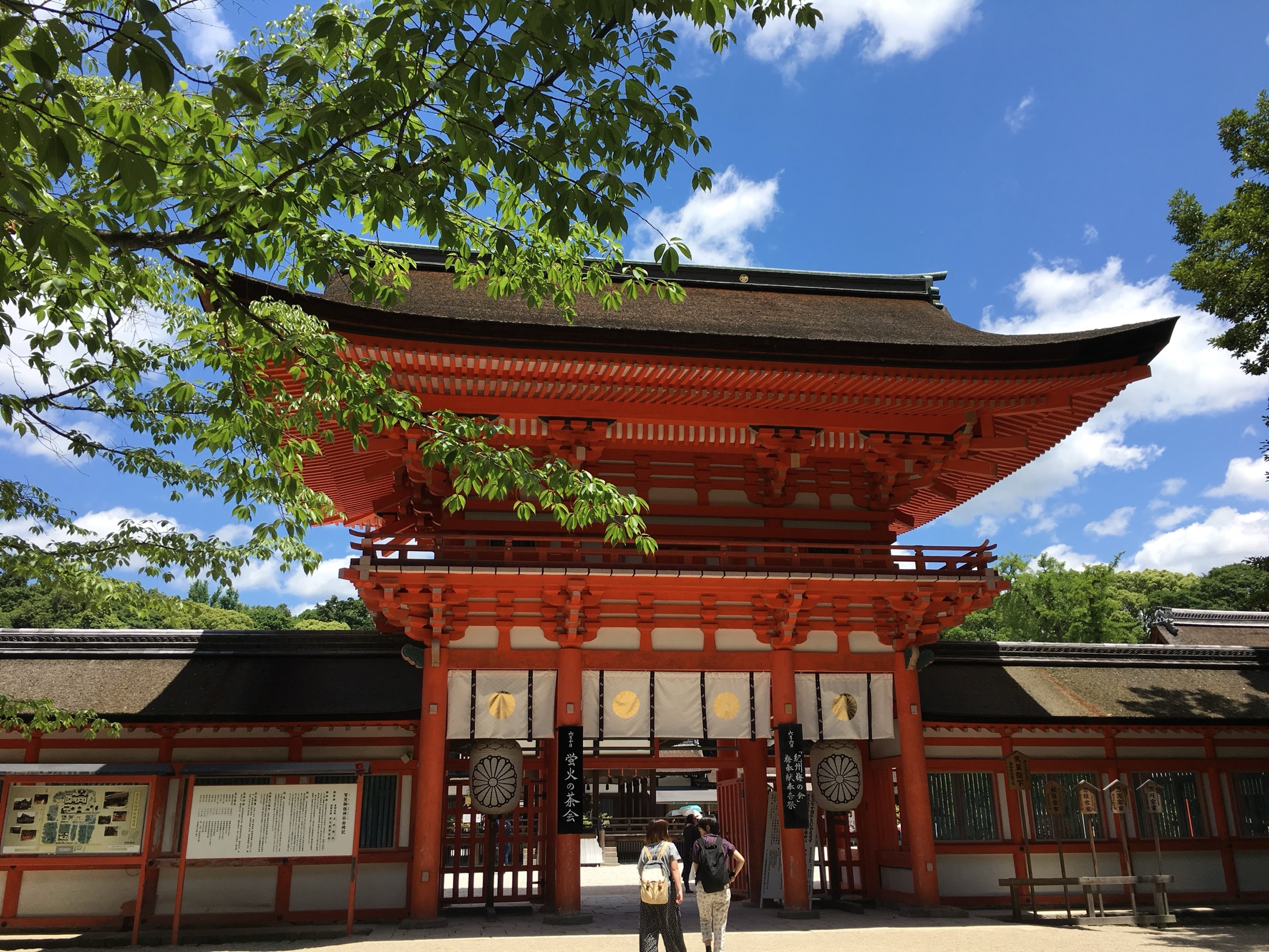 Shimogamo Shrine main Romon gate in vermillion lacquer with curved Japanese tiled roof against a blue summer sky with white clouds, two visitors walking toward the gate, the front courtyard of the UNESCO World Heritage shrine in Kyoto
