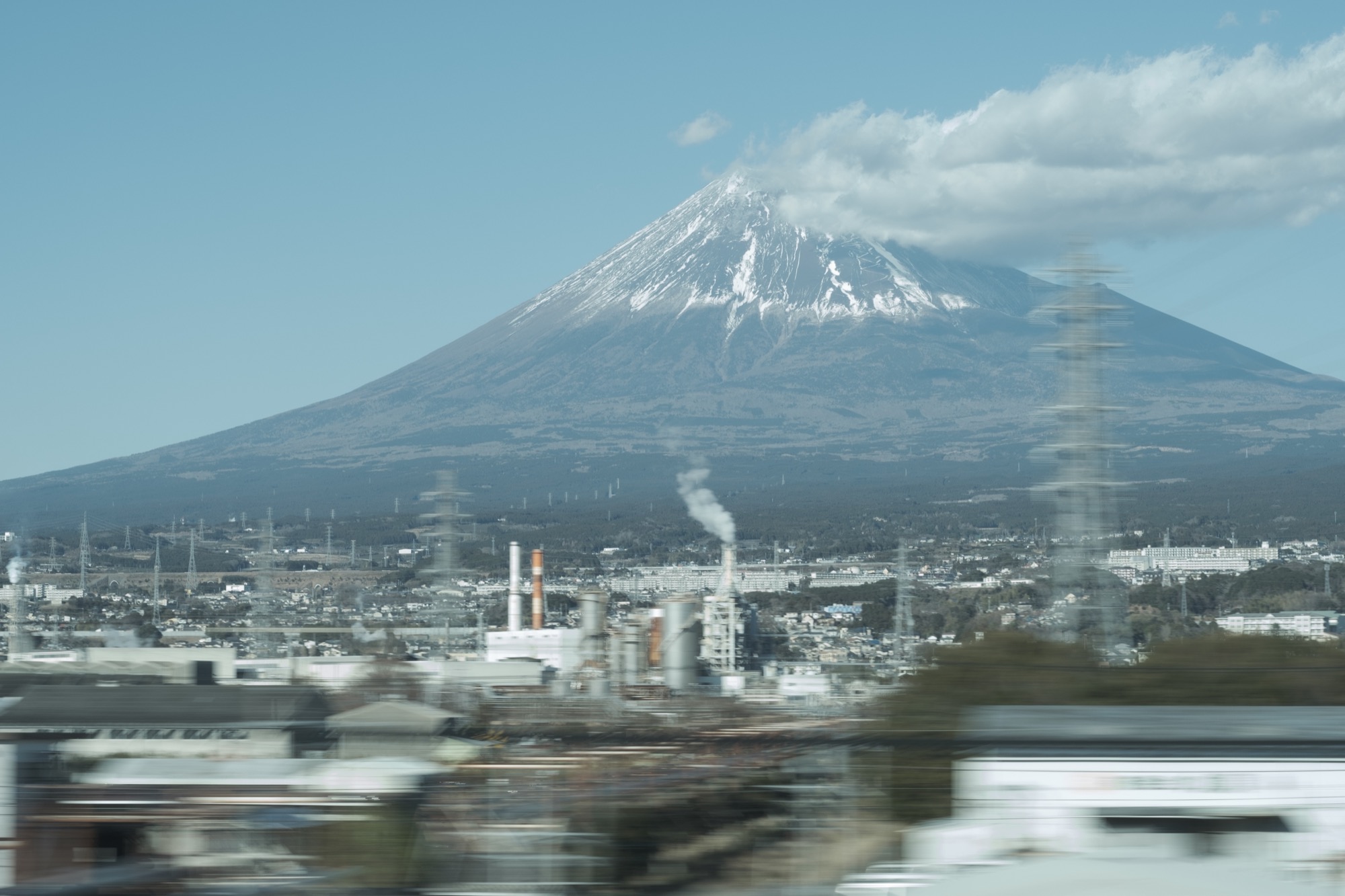 Mt Fuji with snow-capped summit and morning cloud cap photographed from the window of a Tokaido Shinkansen bullet train near Shin-Fuji station with the industrial Fuji City skyline in the foreground and slight motion blur