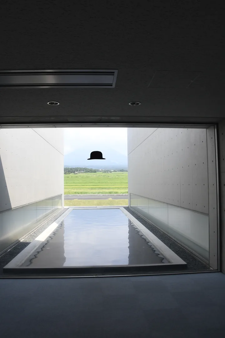 Outdoor passage at Shoji Ueda Museum with a black bowler hat sculpture installation referencing Ueda's signature dune fashion photographs against the rice paddy and Mt Daisen view