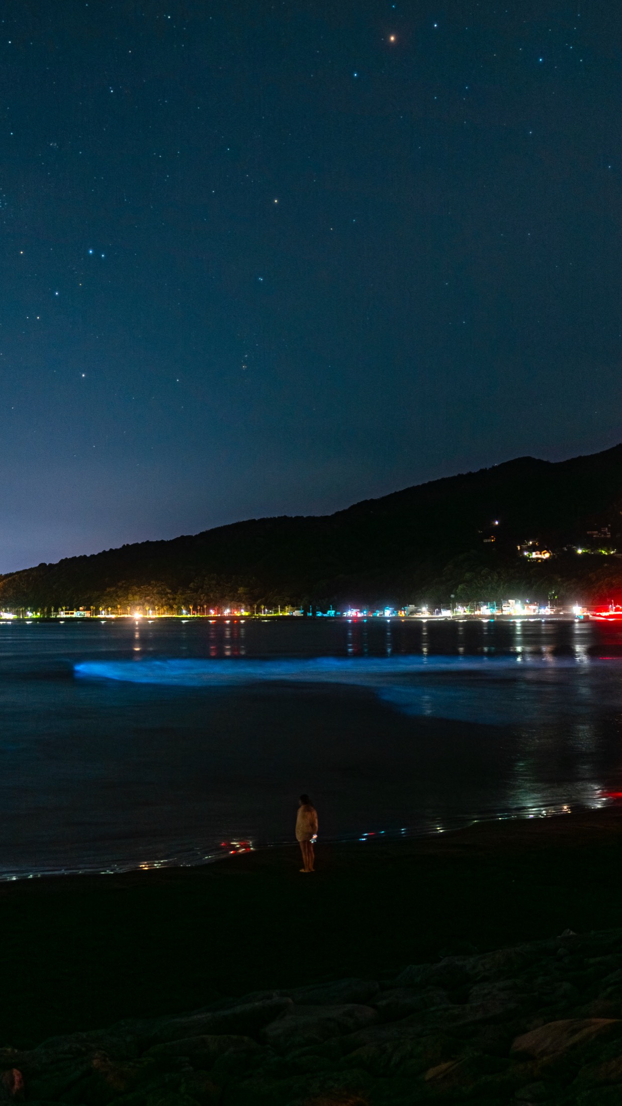 Wide view of Usami Beach in Ito Shizuoka at night with blue bioluminescent waves crashing on shore under a starry sky, town lights across the bay and a lone figure standing on the dark beach