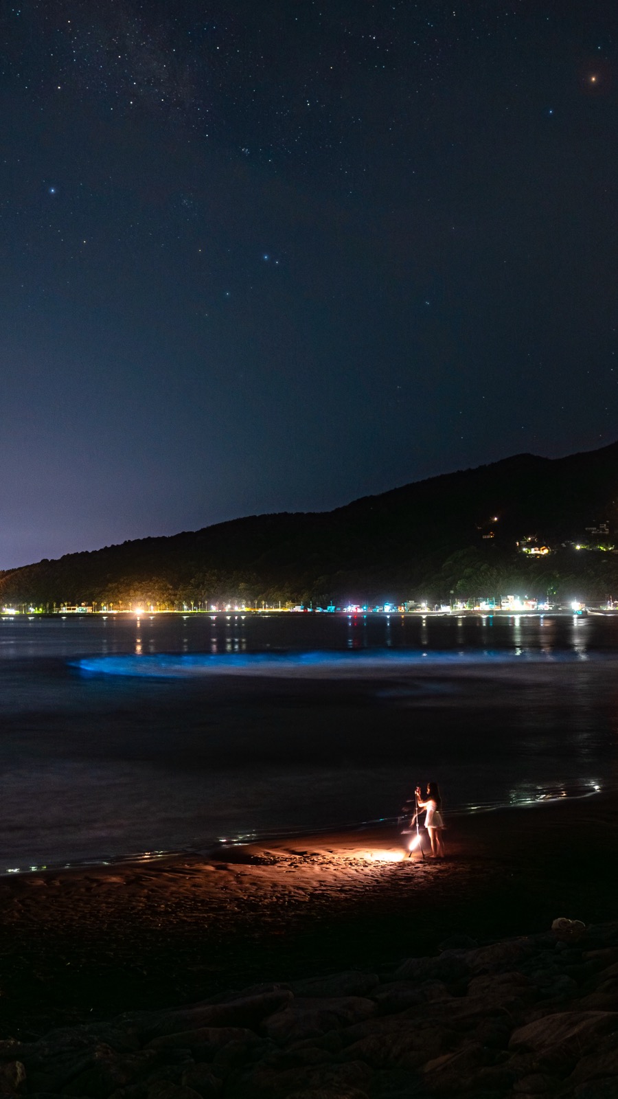 Two people standing on Usami Beach with a glowing lantern as bioluminescent noctiluca waves break behind them and stars fill the night sky over the bay