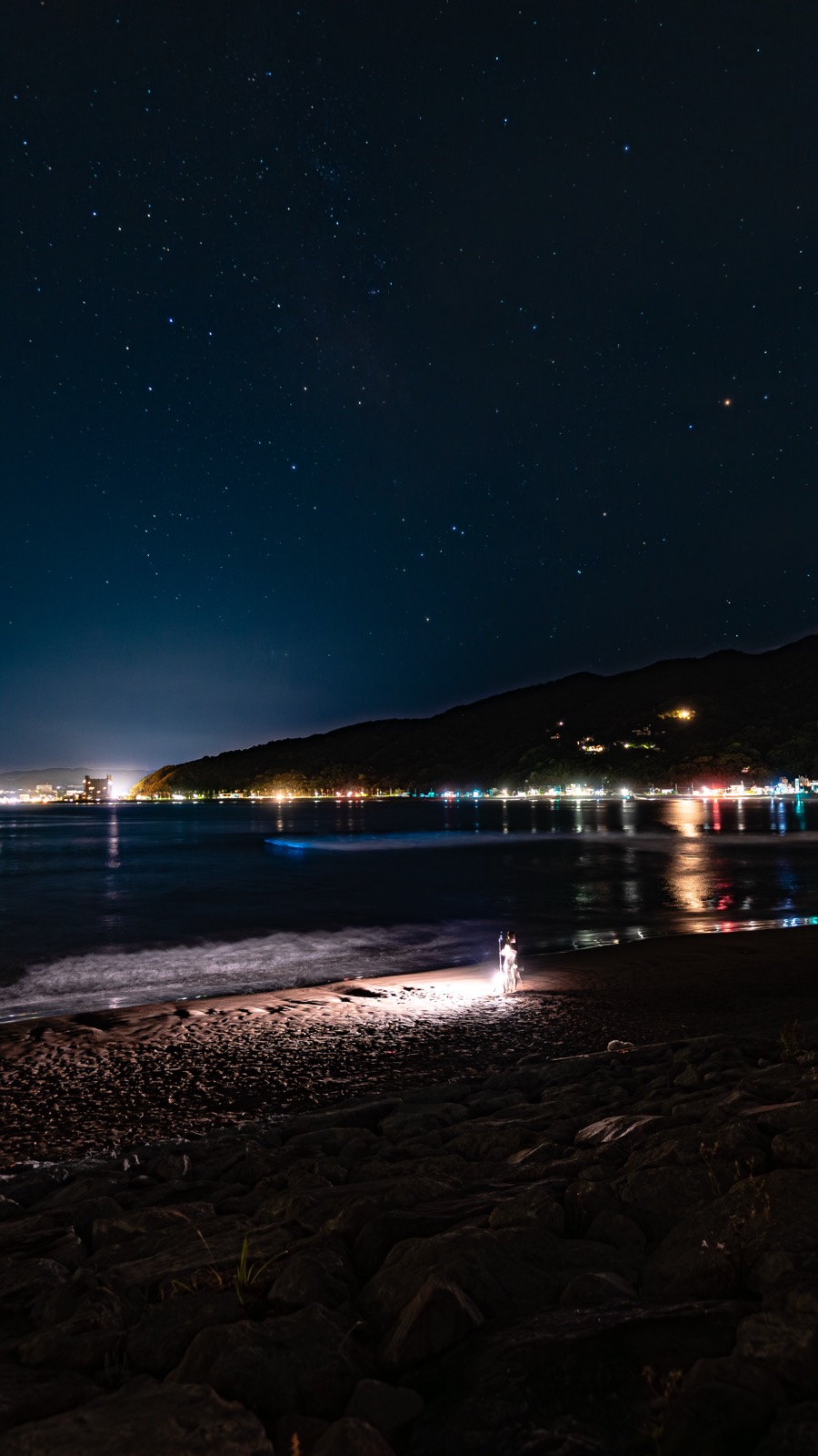 Distant figure with a small lantern on Usami Beach beside the soft blue glow of noctiluca waves with a dense scatter of stars above