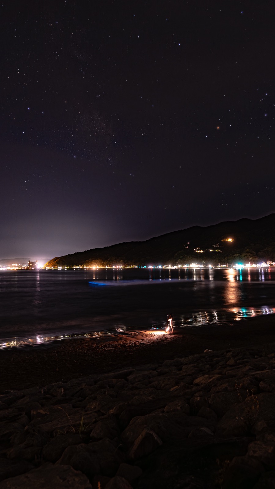 Vertical night view of Usami Beach with a person sitting near a small lantern beside glowing blue noctiluca waves while town lights twinkle across the bay and stars fill the sky