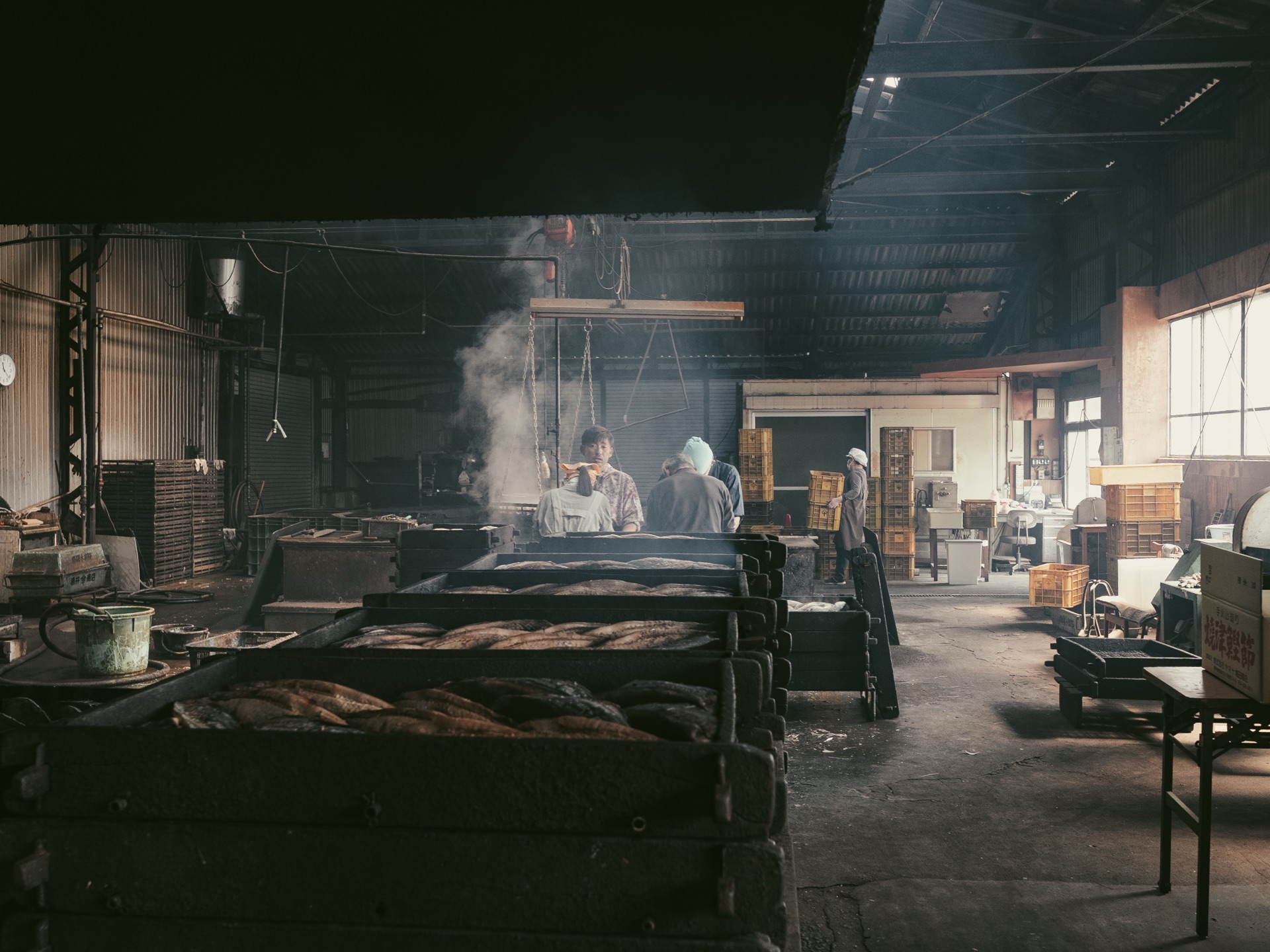 Long wooden trays of smoking bonito loins extending into the dark workshop with two craftsmen at the back
