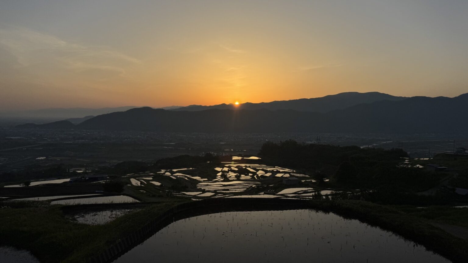 The low sun sitting on a distant ridgeline at sunrise beyond the Obasute rice terraces, the foreground flooded paddies holding a calm sheet of reflected orange and grey dawn sky