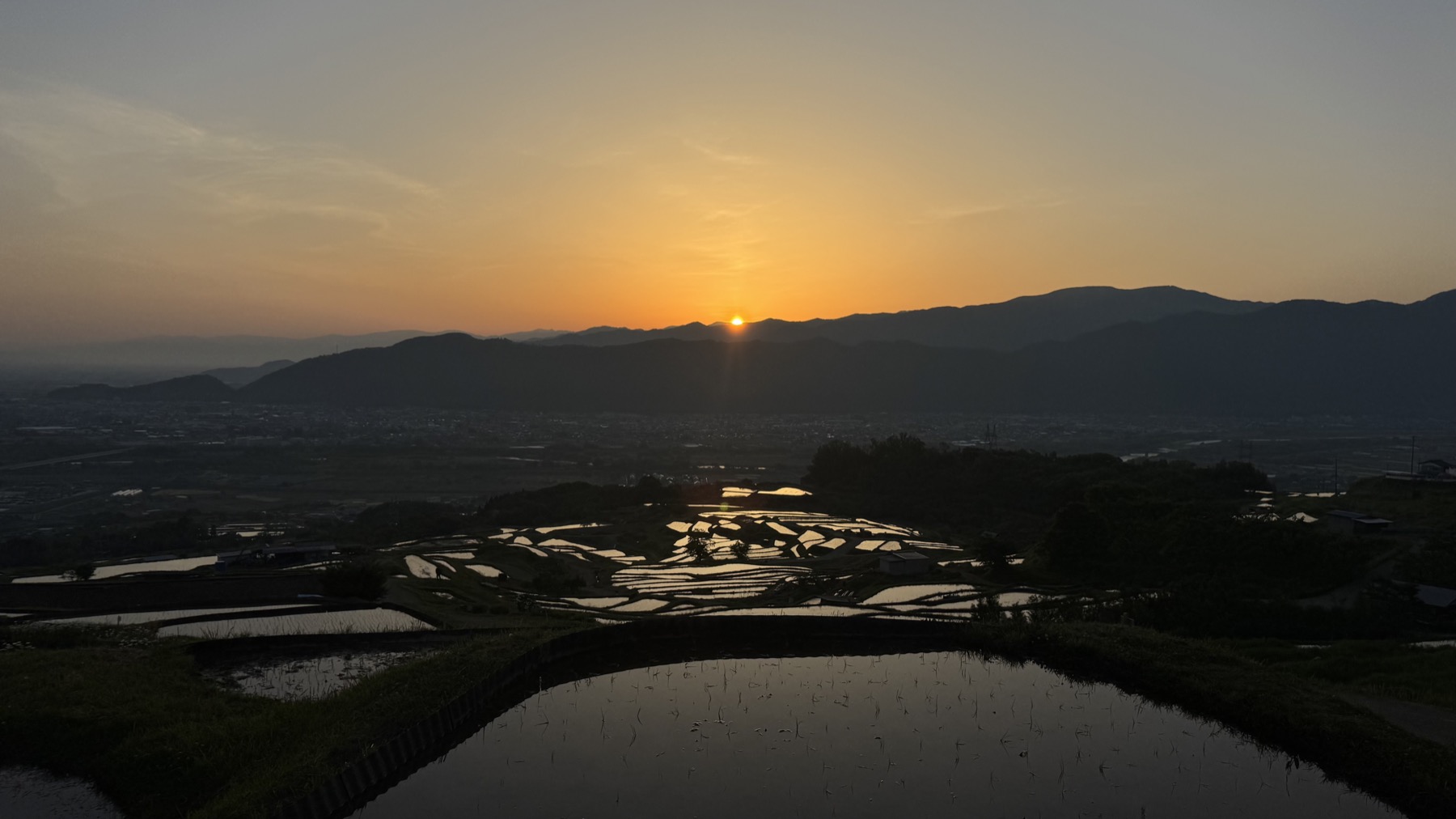 The low sun sitting on a distant ridgeline at sunrise beyond the Obasute rice terraces, the foreground flooded paddies holding a calm sheet of reflected orange and grey dawn sky