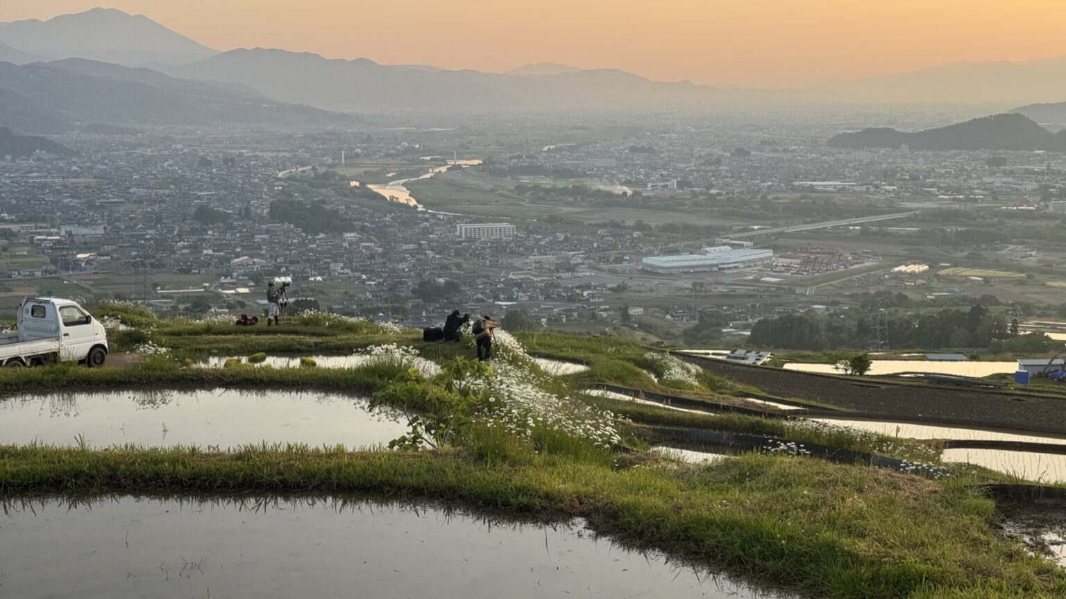 Photographers and a small white farm kei-truck beside the flooded Obasute terraces at sunrise, white wildflowers on the grassy ridges and the city of Chikuma in the early haze below along the Chikuma River