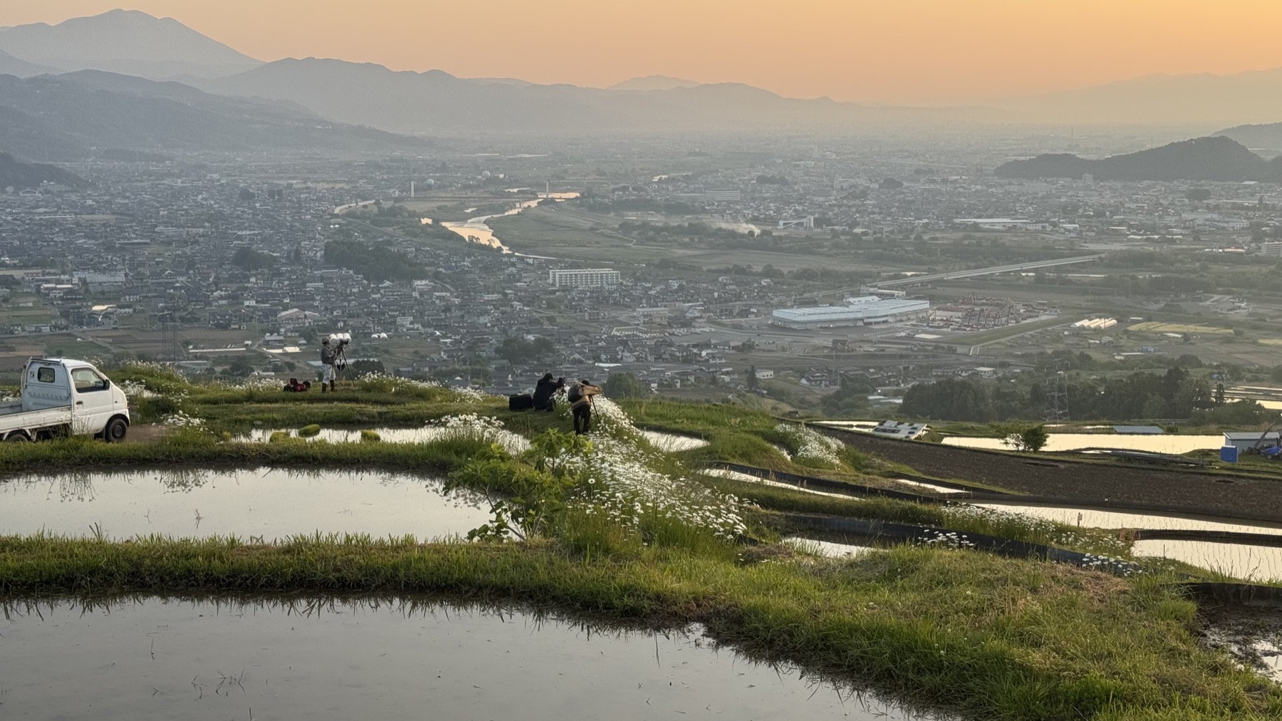 Photographers and a small white farm kei-truck beside the flooded Obasute terraces at sunrise, white wildflowers on the grassy ridges and the city of Chikuma in the early haze below along the Chikuma River