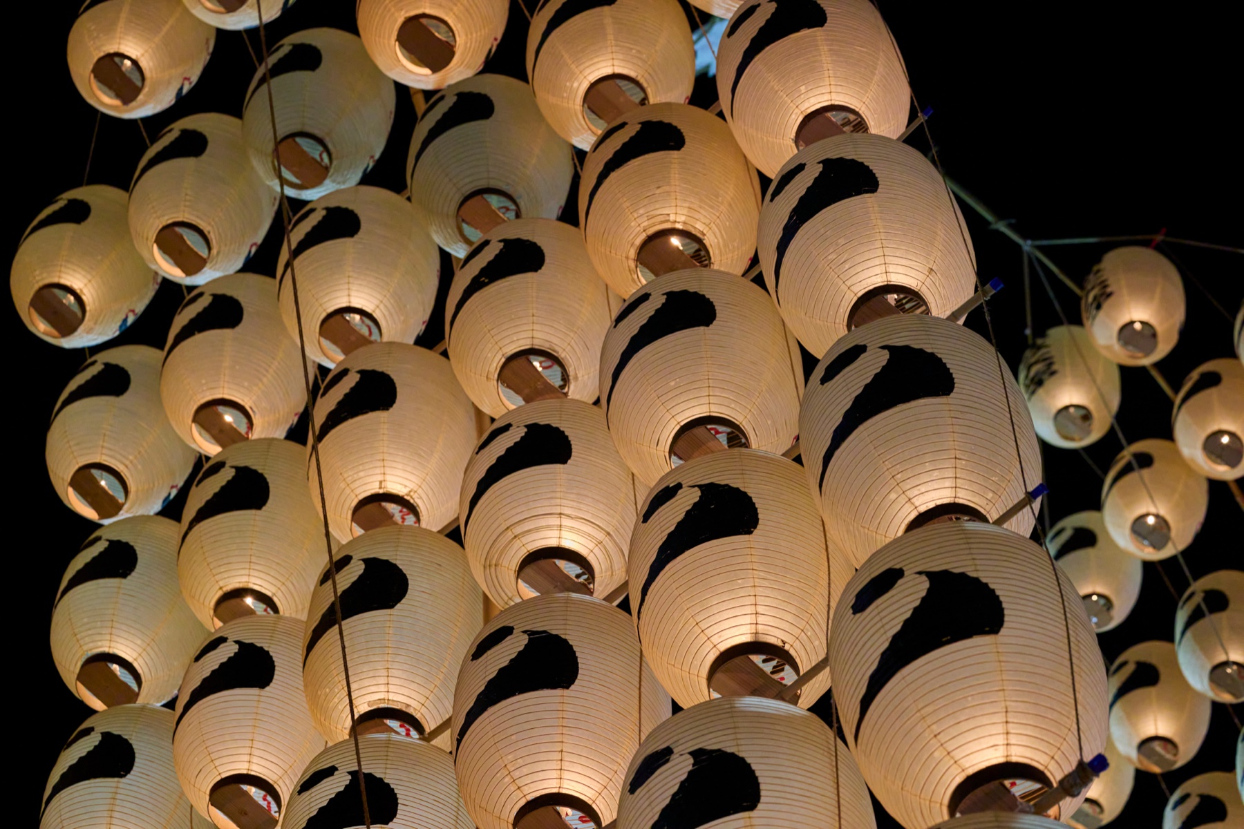 A close-up from below of a kanto pole's grid of round white paper lanterns, each marked with a black neighbourhood crest, glowing against the night sky at the Akita Kanto Festival.