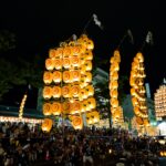 Several tall kanto poles strung with glowing paper lanterns rise above a large nighttime crowd on Kanto Odori during the Akita Kanto Festival, with performers in happi coats balancing the poles in Akita City.