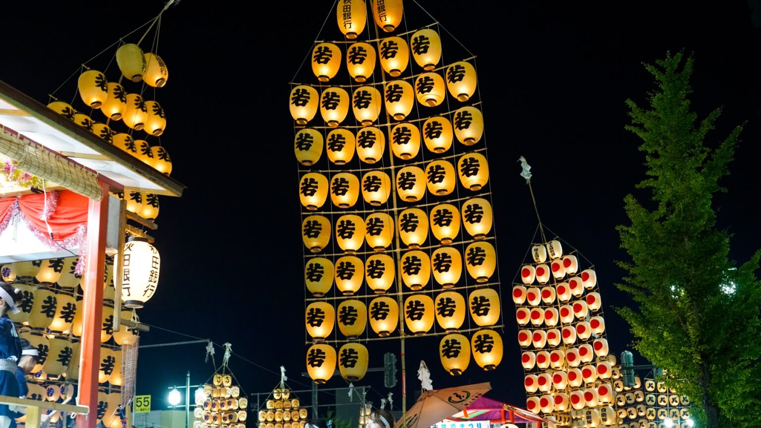 Tall vertical kanto poles covered in lit paper lanterns stand among festival food stalls and a ginkgo tree at night during the Akita Kanto Festival in Akita City.