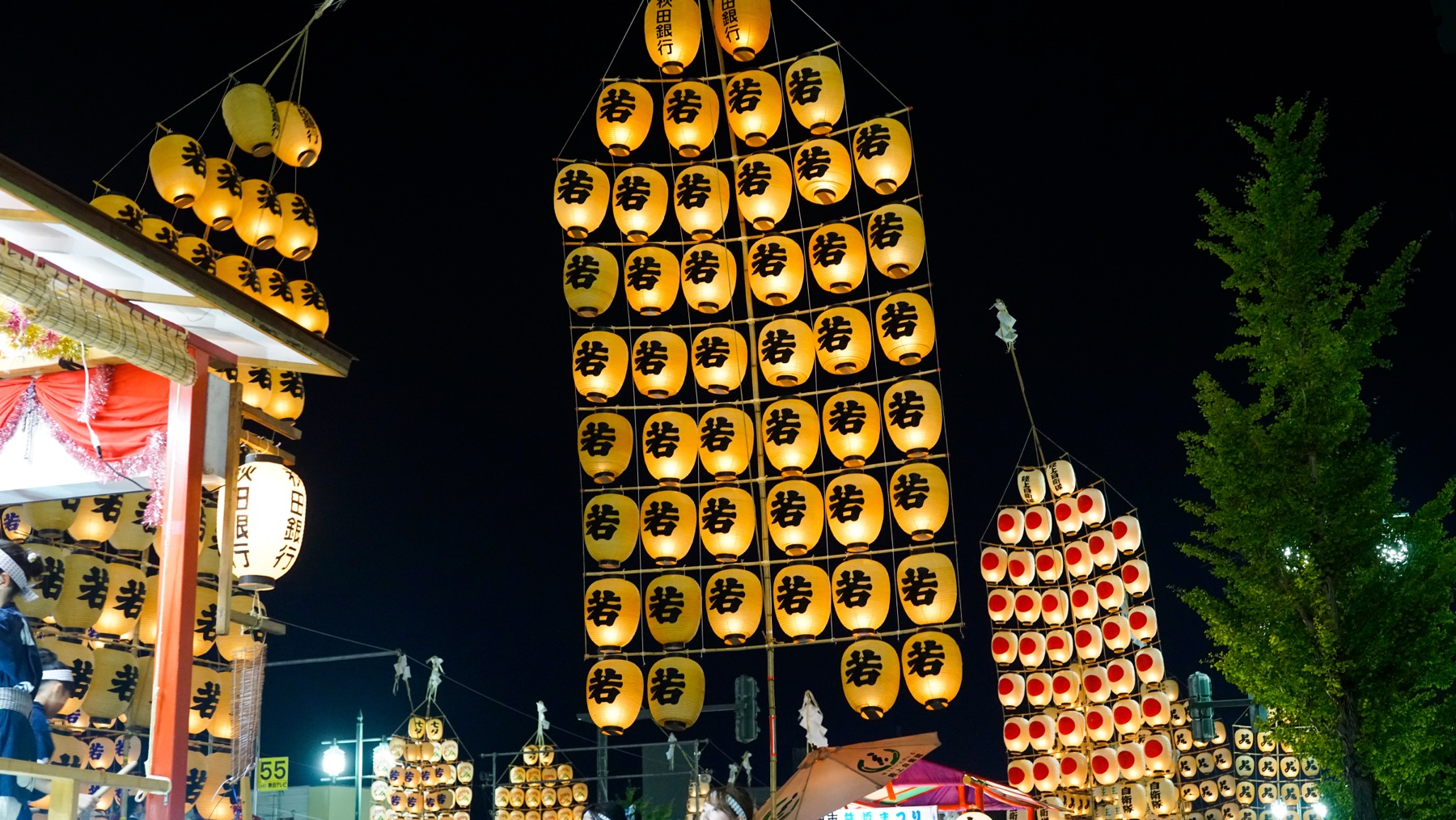 Tall vertical kanto poles covered in lit paper lanterns stand among festival food stalls and a ginkgo tree at night during the Akita Kanto Festival in Akita City.