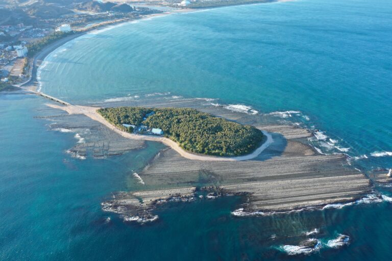 An aerial view of forested Aoshima island off the Miyazaki coast ringed by the concentric wave-cut rock terraces known as the Devils Washboard, with a causeway to the sandy mainland beach and teal sea.