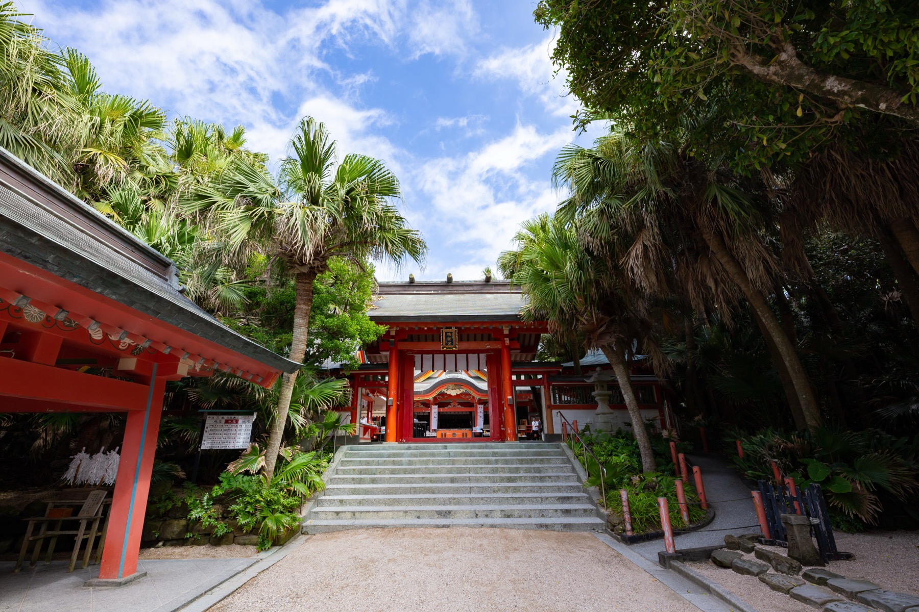 The vermilion gate of Aoshima Shrine at the top of stone steps flanked by subtropical biro palm trees under a blue sky on Aoshima island, Miyazaki.