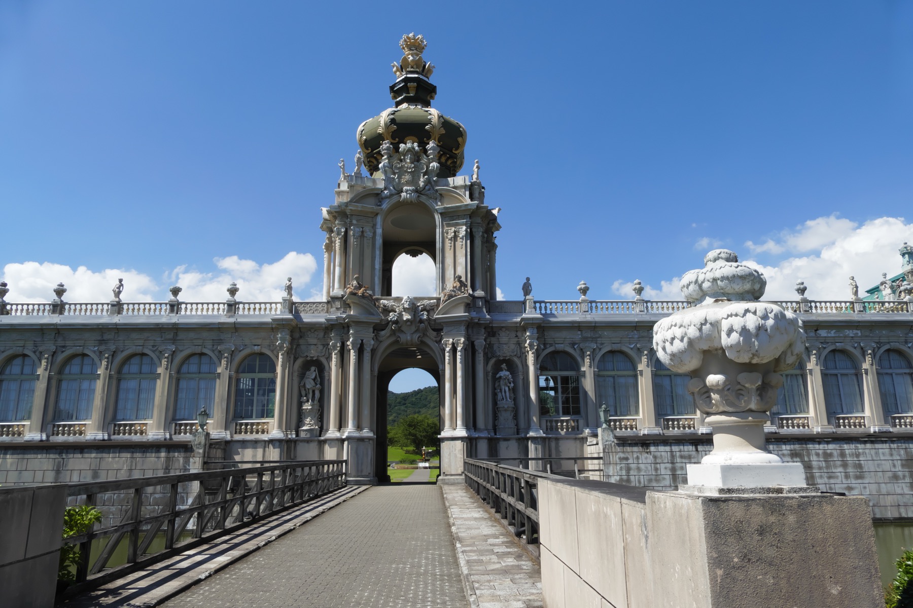 The Kronentor crown gate of the Zwinger Palace replica at Arita Porcelain Park in Saga, a baroque stone archway topped with a gilded crown, with a porcelain urn and a paved walkway leading toward it under a clear blue sky.
