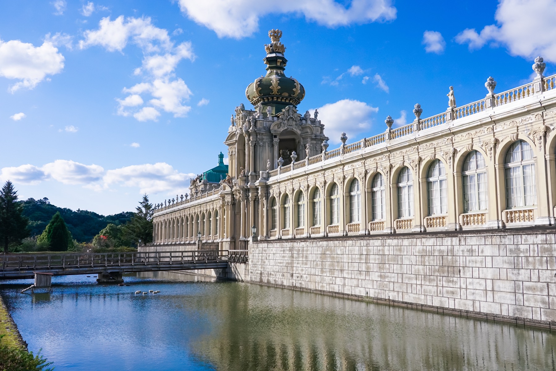 The long baroque palace wing and golden crowned dome of the Zwinger replica at Arita Porcelain Park in Saga, lined with arched windows and stone statues, mirrored in the still canal running alongside it.