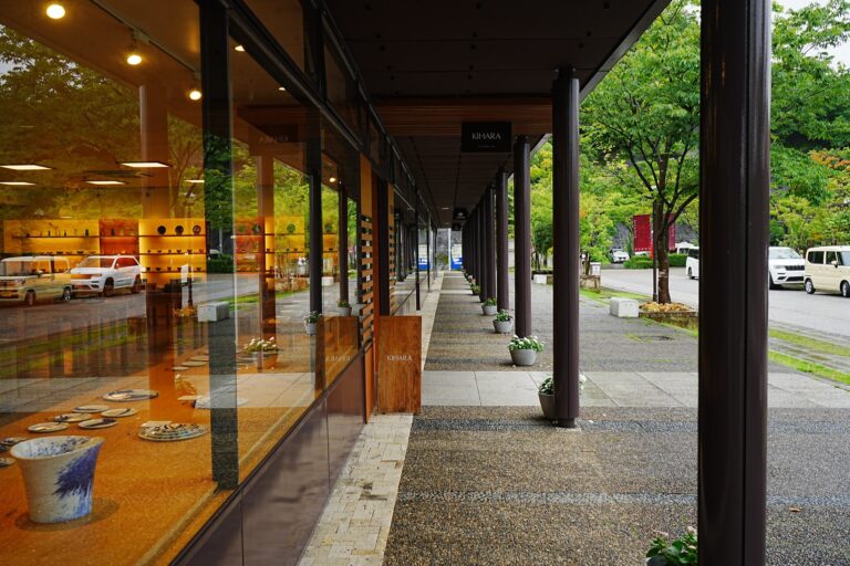 The long covered shopping arcade of Arita Sera in Arita, Saga, glass-fronted porcelain shops with displays of plates and vases on one side and a paved walkway with planters and trees on the other