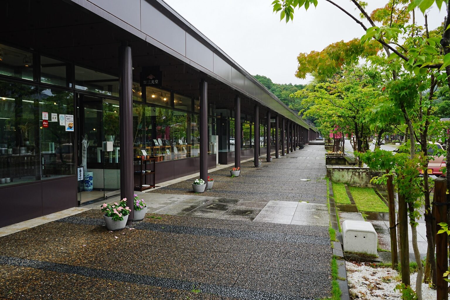 The covered walkway of the Arita Sera porcelain complex on a rainy day, a long row of glass-fronted ceramic shops with potted flowers along the wet paving, Arita, Saga