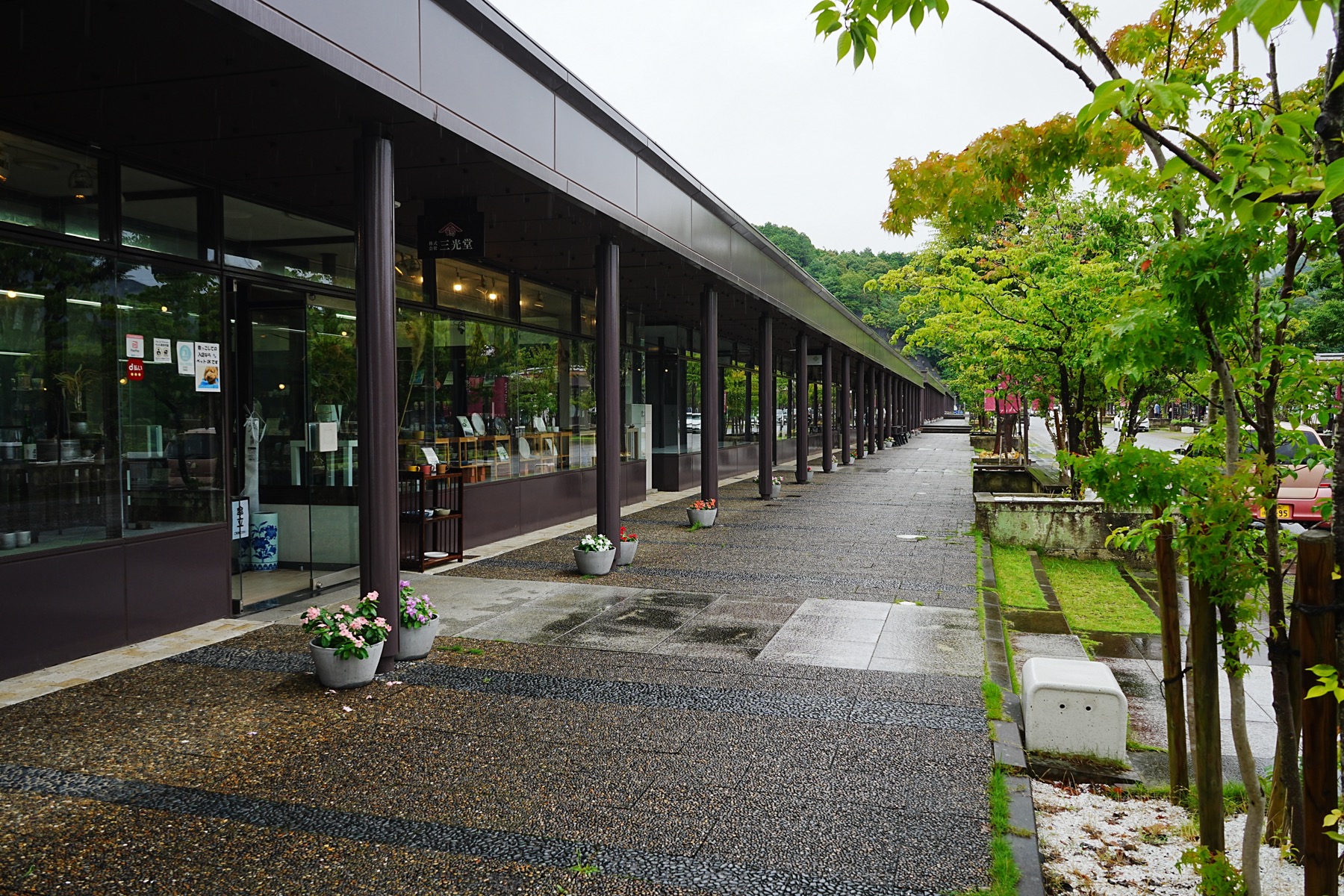 The covered walkway of the Arita Sera porcelain complex on a rainy day, a long row of glass-fronted ceramic shops with potted flowers along the wet paving, Arita, Saga
