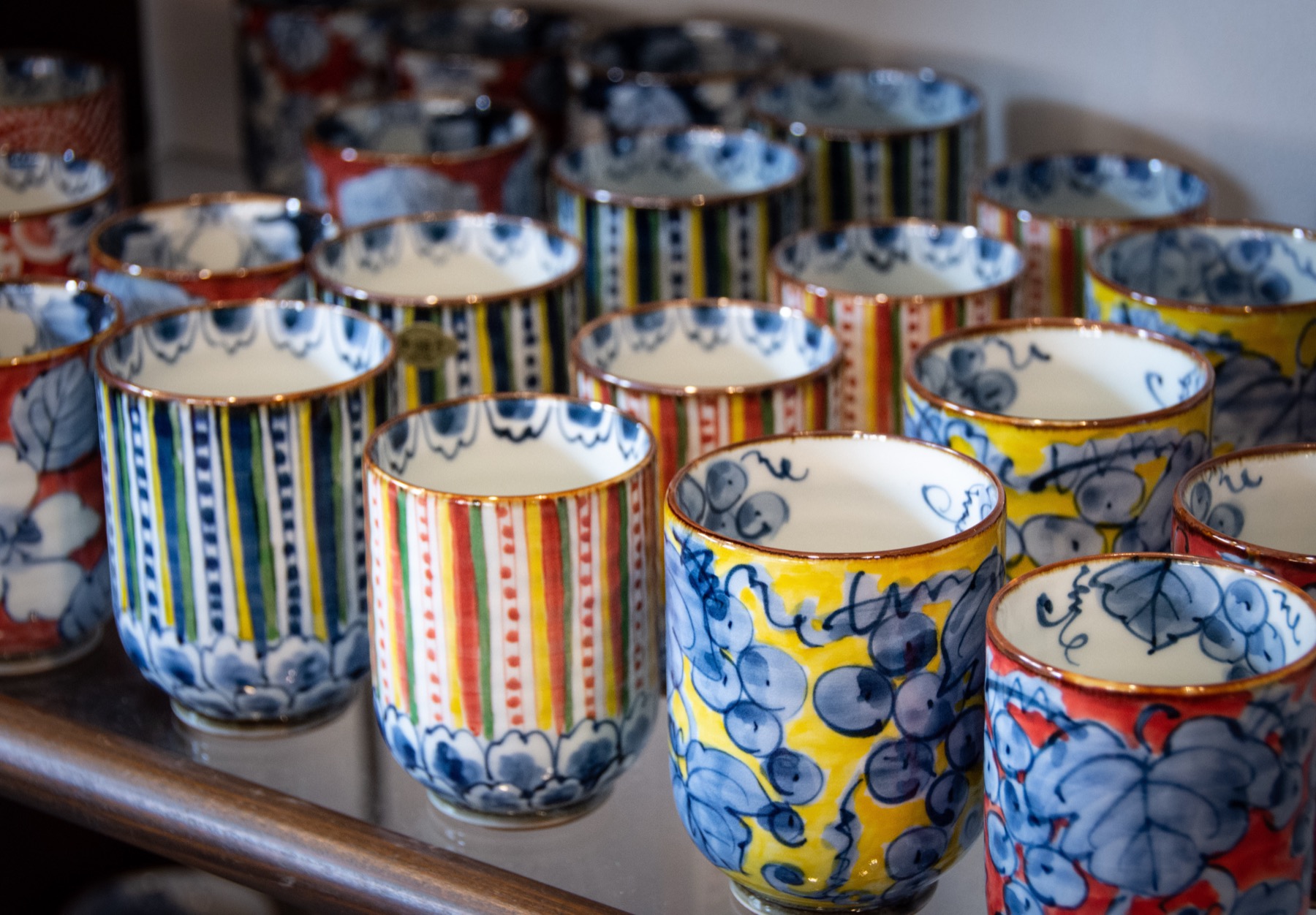 Rows of colourful Arita-ware tea cups (yunomi) on a shop shelf, hand-painted with blue florals, multicoloured stripes and gold rims, on display at a porcelain shop in Arita, Saga