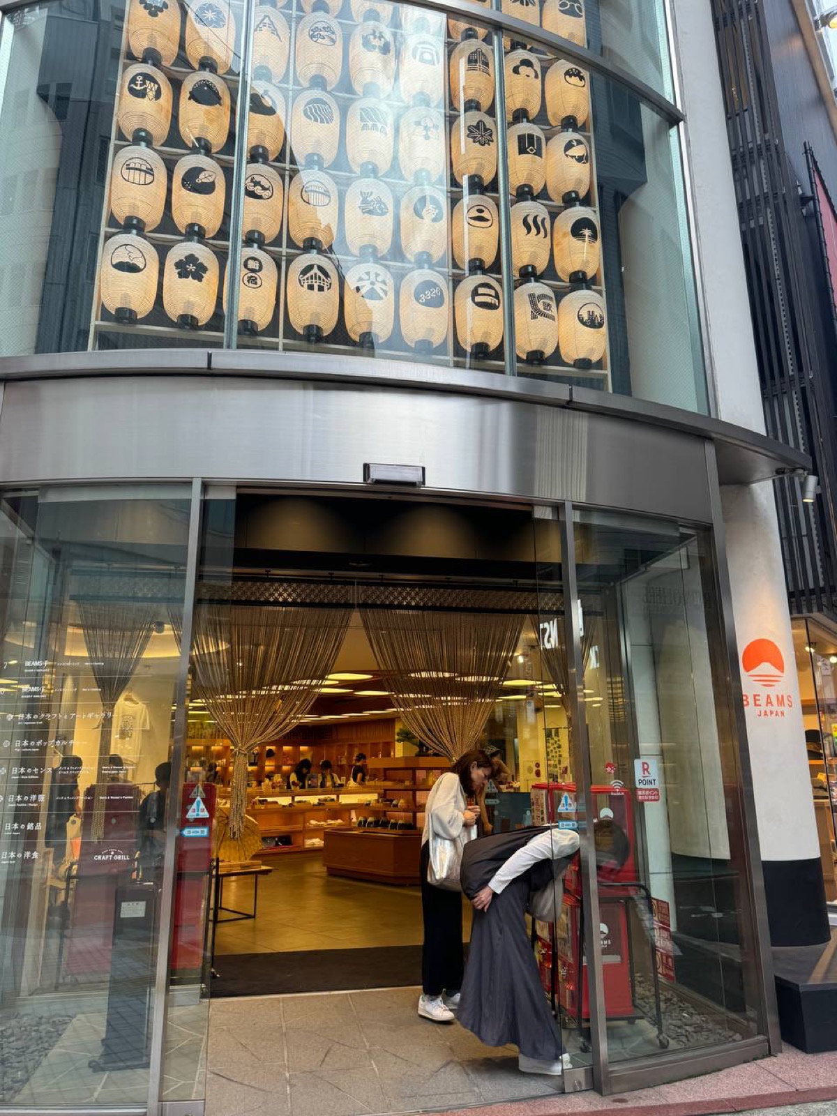 The curved glass revolving-door entrance of BEAMS JAPAN Shinjuku, topped with a display of wooden panels painted with Japanese motifs like Mt Fuji and plum blossoms, the wood-shelved interior visible inside.