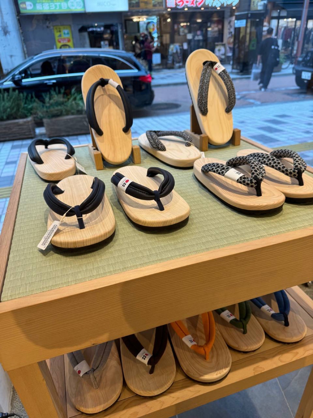A display of wooden geta and setta sandals with black, patterned and coloured thong straps arranged on a tatami-topped wooden stand at BEAMS JAPAN Shinjuku, the Shinjuku street visible through the window.