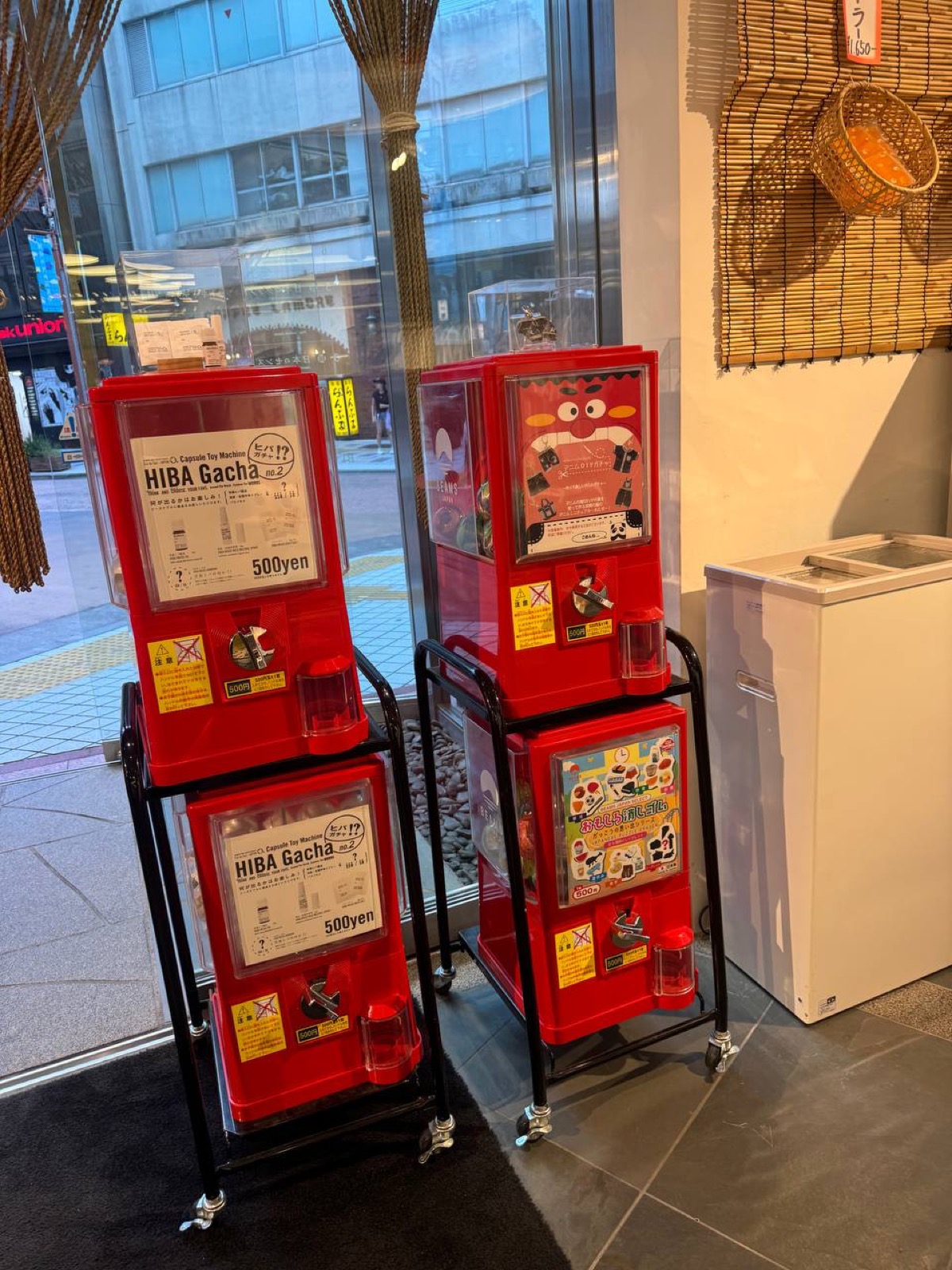 Red capsule-toy gachapon machines labelled 'HIBA Gacha 500 yen' standing by the window inside BEAMS JAPAN Shinjuku, with a woven bamboo basket hung on the wall beside them.