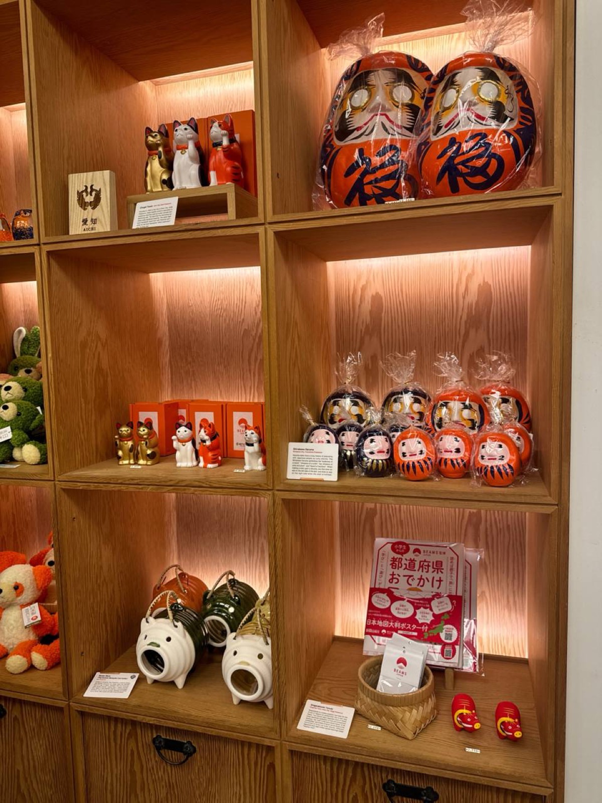 Wooden cube shelves at BEAMS JAPAN Shinjuku displaying maneki-neko beckoning-cat figurines, large red fuku daruma dolls, small daruma sets, plush toys and ceramic ornaments, lit with warm shelf lighting.