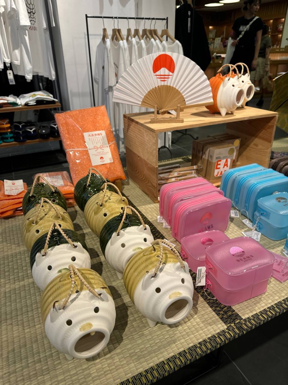 A tatami-topped table at BEAMS JAPAN Shinjuku displaying a folding fan with a rising-sun logo, round white ceramic ornaments with faces, pink and blue plastic bento boxes and folded T-shirts behind.