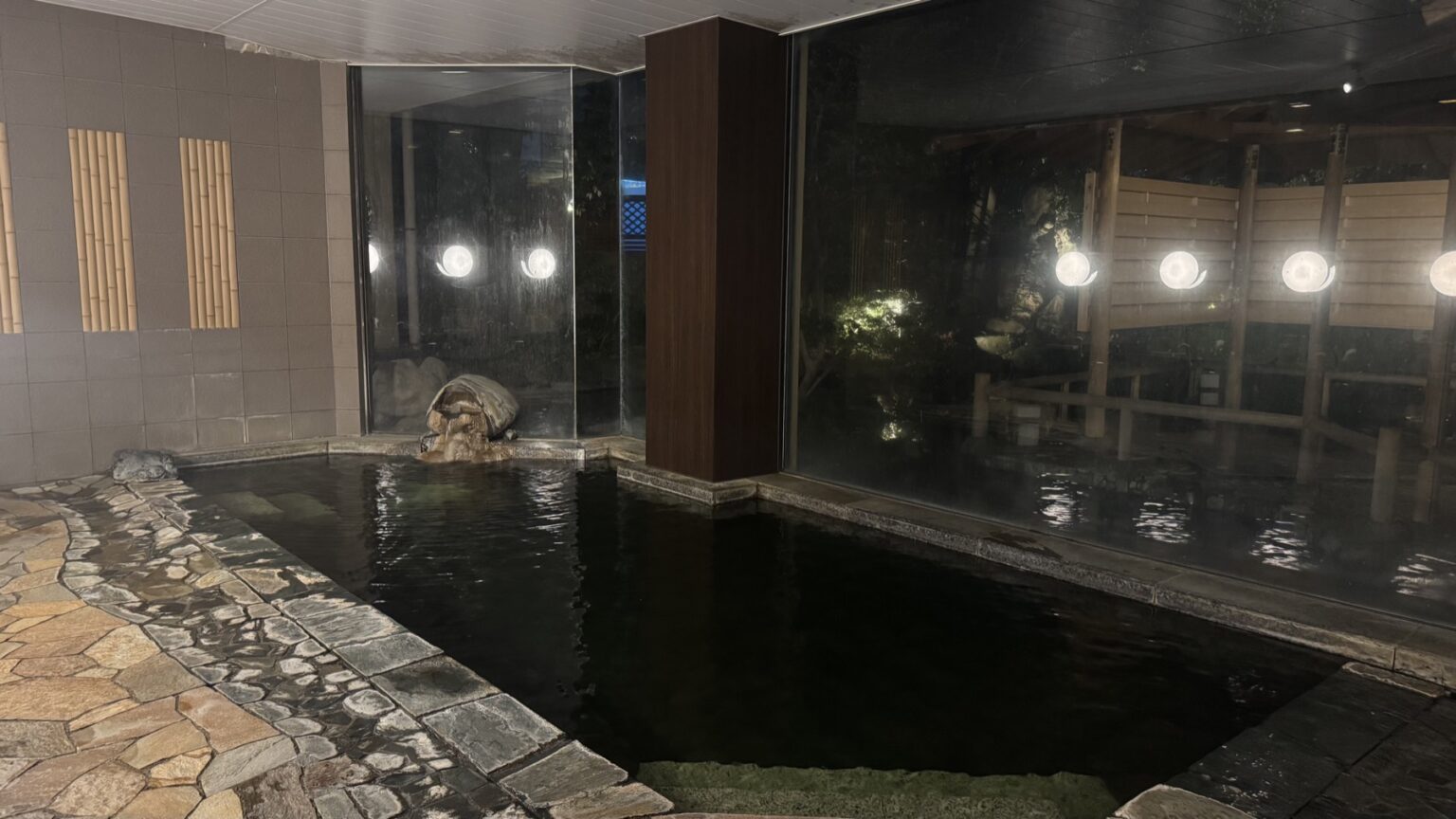 An indoor hot-spring bath at Club Wyndham Chikumakan with a dark stone pool, round wall lights and large windows looking onto an illuminated garden, in Togura-Kamiyamada Onsen, Nagano