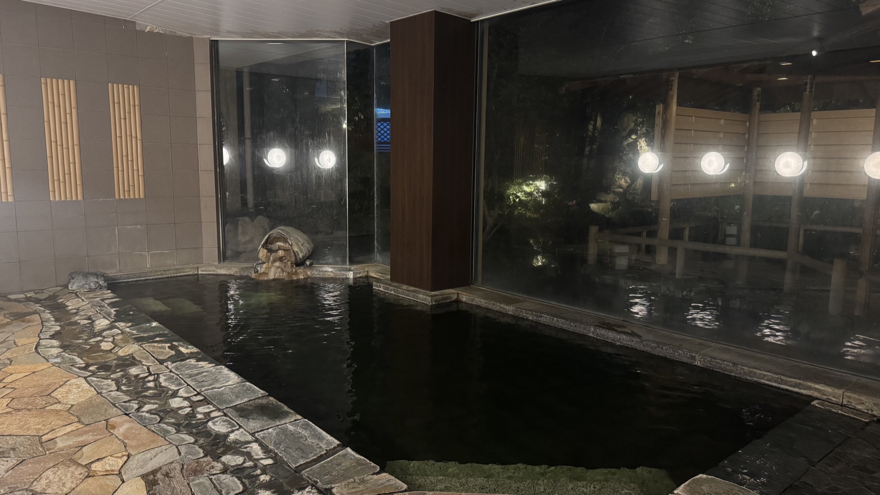 An indoor hot-spring bath at Club Wyndham Chikumakan with a dark stone pool, round wall lights and large windows looking onto an illuminated garden, in Togura-Kamiyamada Onsen, Nagano