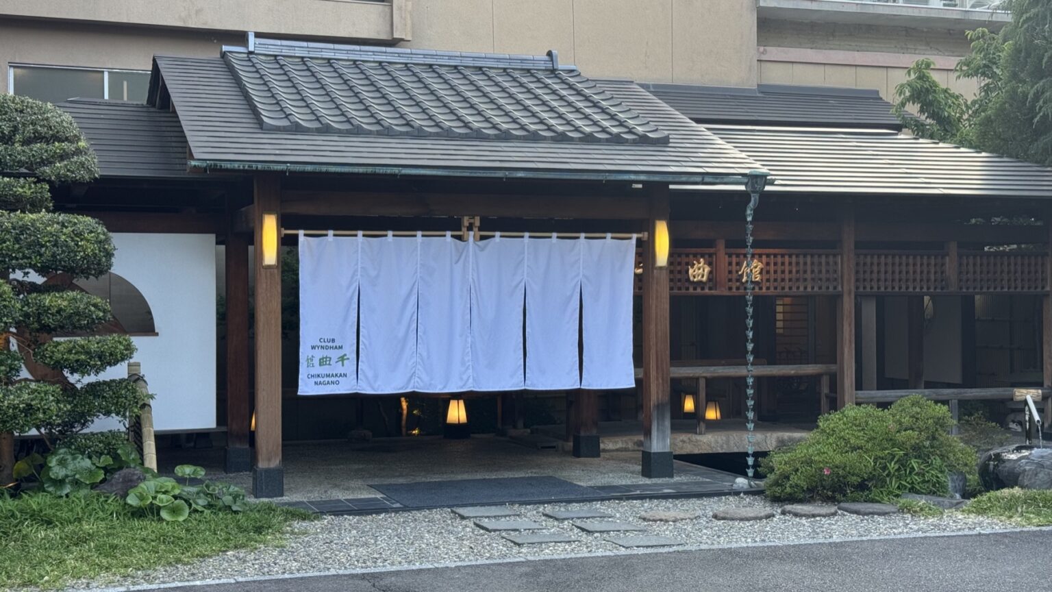 The traditional tiled entrance gate of Club Wyndham Chikumakan in Togura-Kamiyamada Onsen, with a white noren curtain reading Club Wyndham Chikumakan Nagano, lanterns and a stone path through a small garden