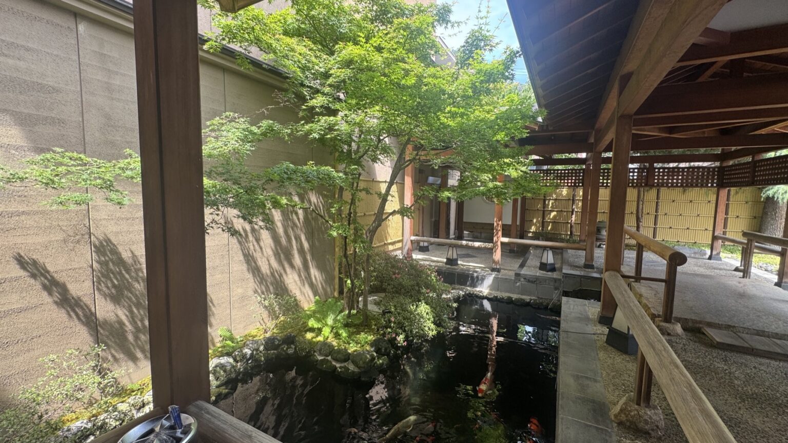 A green inner courtyard at Club Wyndham Chikumakan in daylight, a maple tree over a koi pond with carp, wooden walkways and the ryokan's tiled eaves, in Togura-Kamiyamada Onsen, Nagano