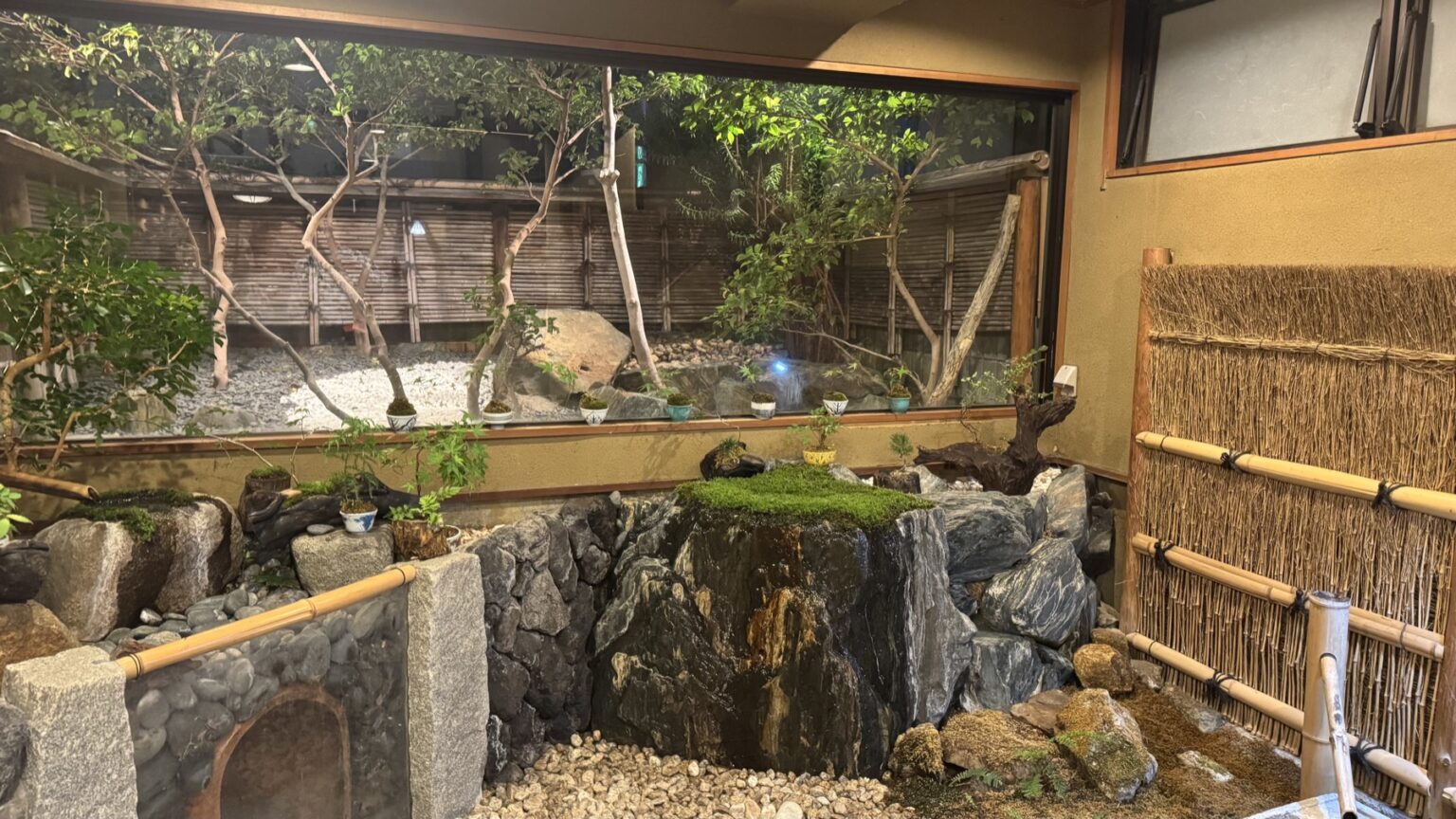 A private reservable bath at Club Wyndham Chikumakan looking through glass onto a rock-and-moss garden with a small stream and a bamboo screen, in Togura-Kamiyamada Onsen, Nagano