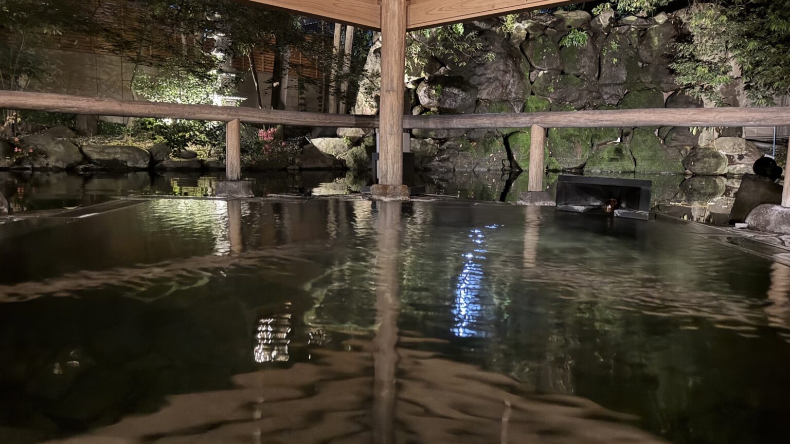 An open-air hot-spring bath at Club Wyndham Chikumakan in Togura-Kamiyamada Onsen at night, the dark water lit softly and reflecting a wooden pavilion and a rock-and-greenery garden, in Chikuma, Nagano