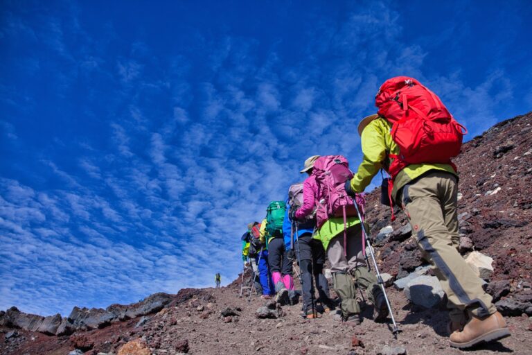 A line of climbers in colourful gear with backpacks and trekking poles ascending the red volcanic gravel slope of Mt Fuji under a deep blue summer sky during the July to September climbing season