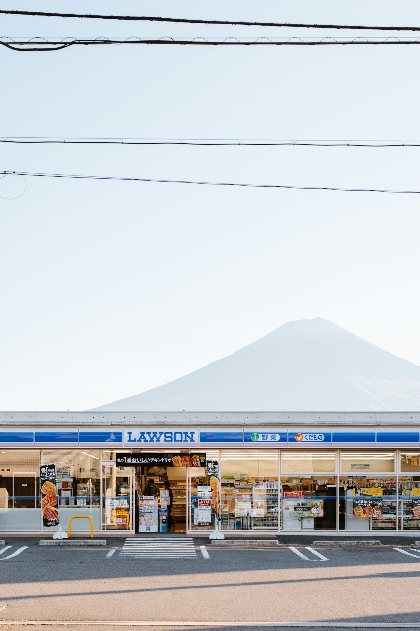 A Lawson convenience store with Mount Fuji rising directly behind it on a clear early-summer morning in the Fuji Five Lakes area of Japan, a quiet car park in front and the snow already gone from the dark summit