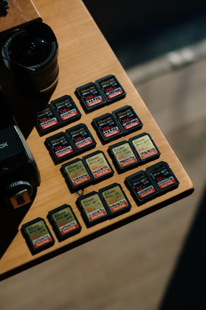About eighteen SanDisk Extreme SD cards from 32GB to 256GB laid out in neat rows on a wooden table beside a camera, the kind of fast-card stack a travel photographer cycles through on long shooting trips.