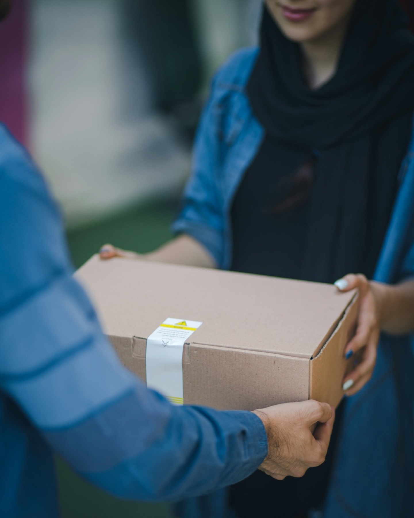 A close-up of a plain brown cardboard parcel being handed over with both hands between two people, one in a denim shirt and headscarf, the kind of hand-off you get at a hotel front desk or a convenience-store counter in Japan