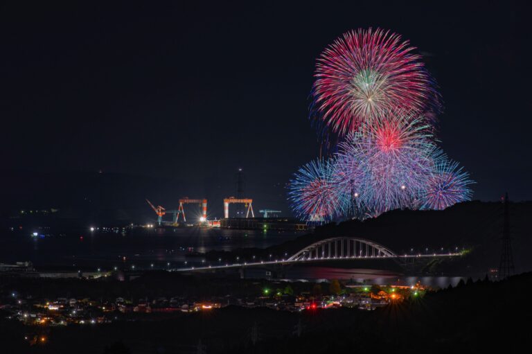 Colorful red, pink and blue fireworks bursting over Imari Bay at night with a lit arch bridge, shipyard cranes and town lights below, at a summer hanabi festival in Saga, Japan
