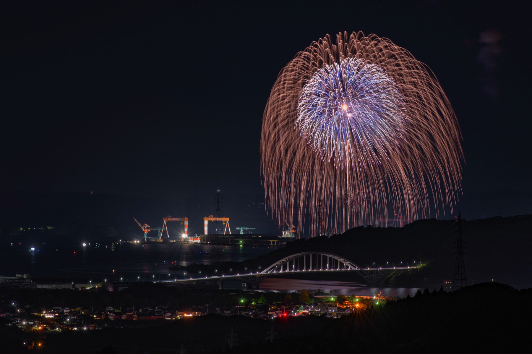 A large golden willow firework opening over Imari Bay at night above a lit arch bridge and harbour lights, at a summer fireworks festival in Saga, Japan